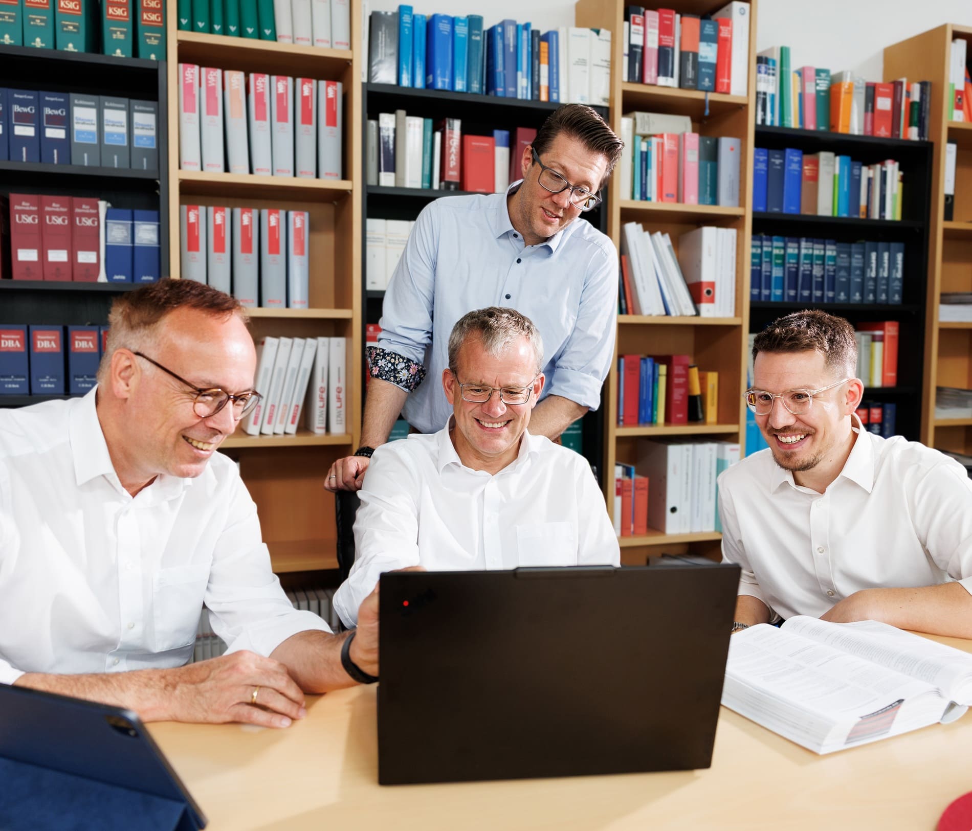 Vier Experten für Datenschutzberatung besprechen Inhalte an einem Laptop vor einem gut gefüllten Bücherregal.